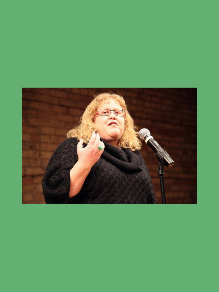Alison Bergblom Johnson, a plus-sized woman with curly blonde hair wearing a black sweater speaking into a silver and black microphone